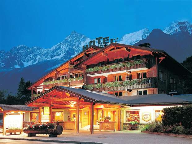 A wooden chalet-style hotel with bright lights illuminates the evening, surrounded by mountainous scenery. "HOTEL" is displayed prominently on the roof, with balconies adorned with flower boxes.